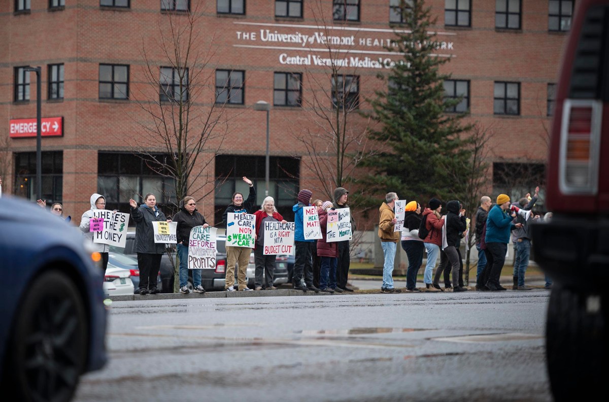 A group of people protest outside a medical facility, holding signs about health care and profit. Some signs are counted "Healthcare not profit" and "We can't pay for the job.