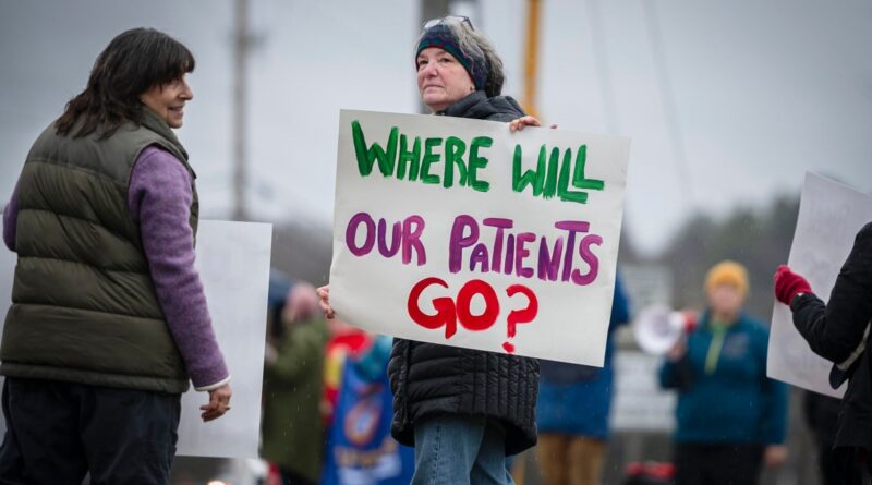 A person is holding a reading sign "Where will our patients go?" during external protests and others nearby.