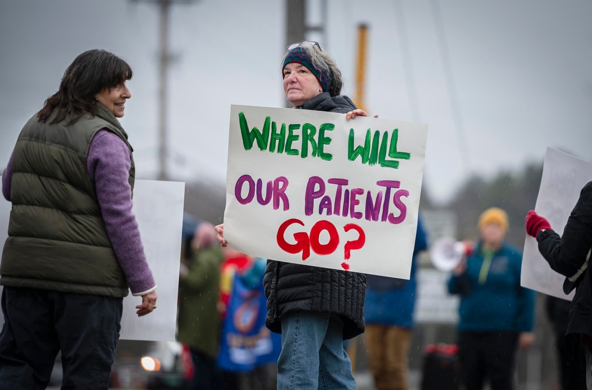 A person is holding a reading sign "Where will our patients go?" during external protests and others nearby.